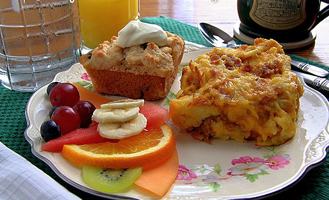 A plated hot breakfast: egg and bread pudding, muffins, a fruit platter, orange juice and coffee